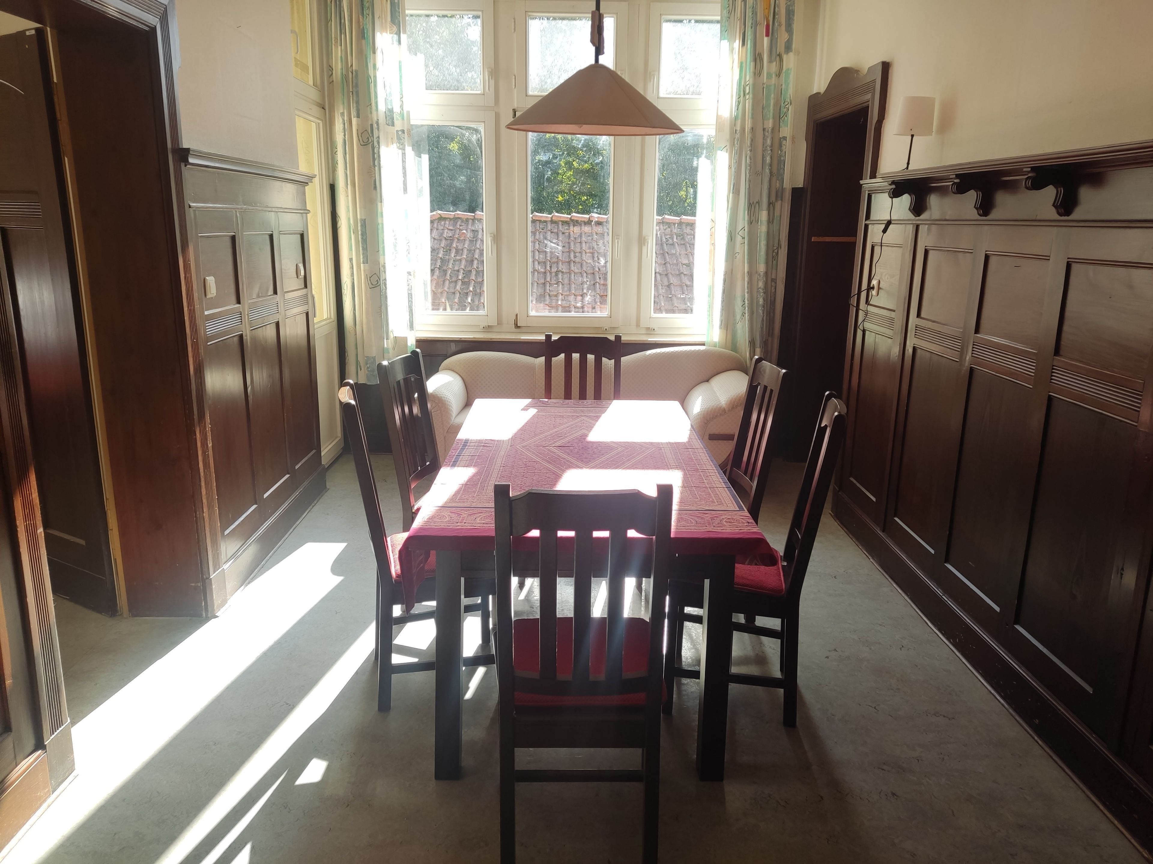 A dining area with a table set for six, surrounded by wooden chairs, a hanging light fixture, and a view through large windows.