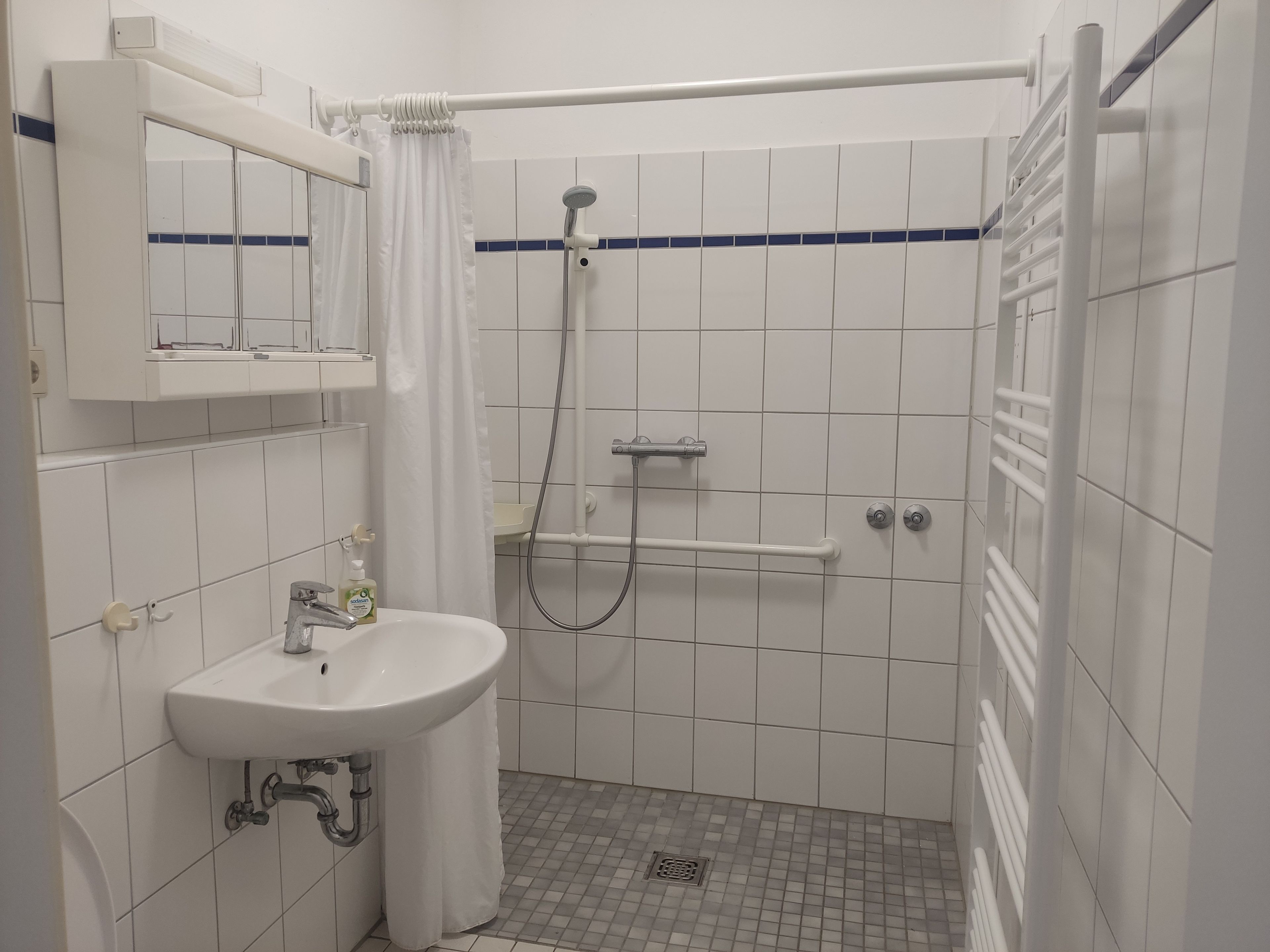 Ein kleines, weißes Badezimmer mit Waschbecken, Dusche und Spiegel.