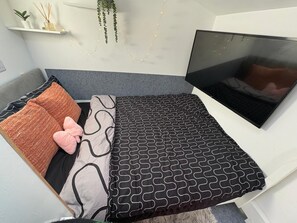 Kompakti sänky, jossa on harmaa pääty, musta kuviollinen päiväpeite ja yksi vaaleanpunainen koristetyyny.