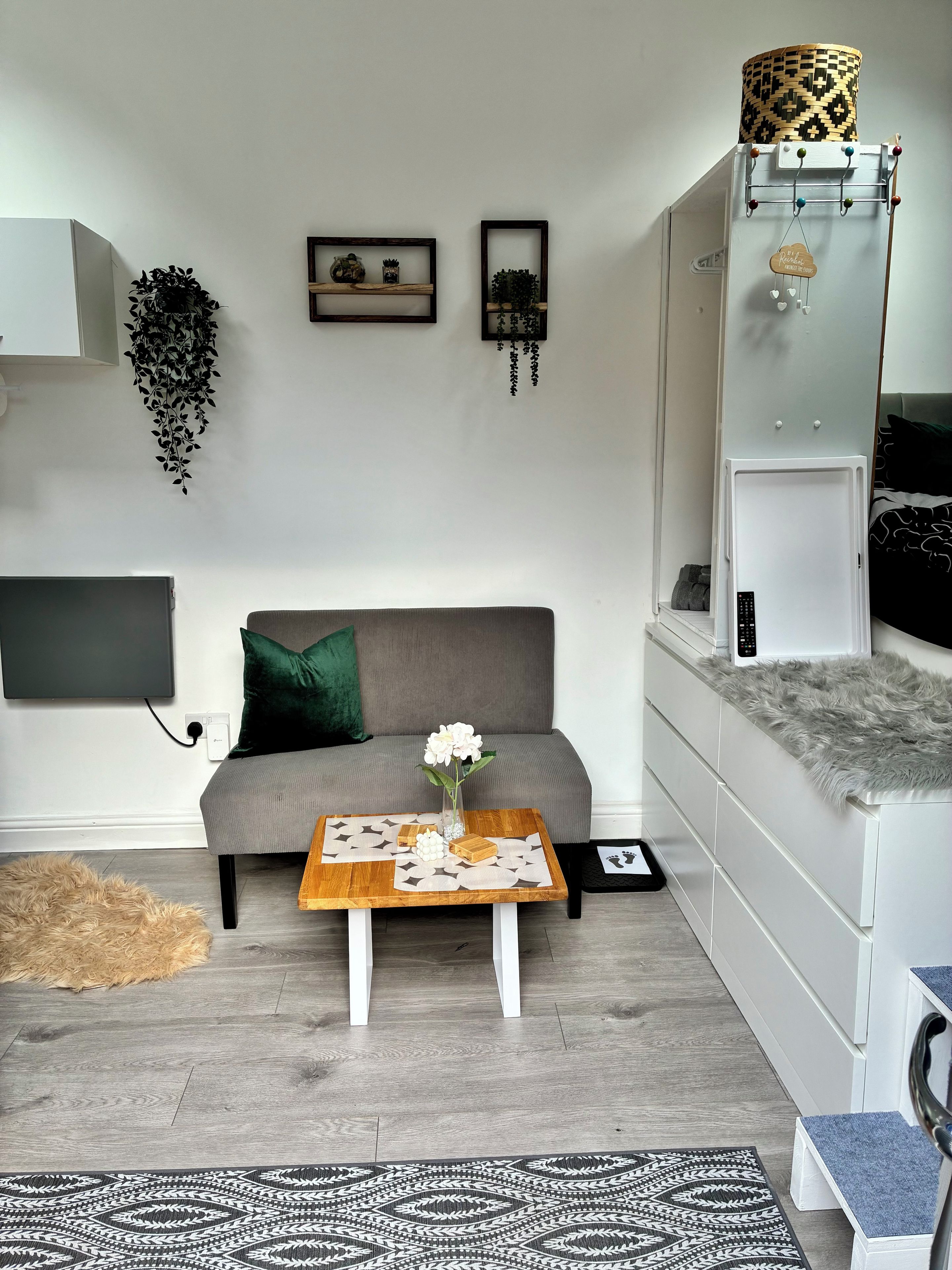 Una sala moderna con un sofá gris, una mesa de centro de madera con objetos decorativos, un televisor montado en la pared y un mueble blanco con un tapete estampado.