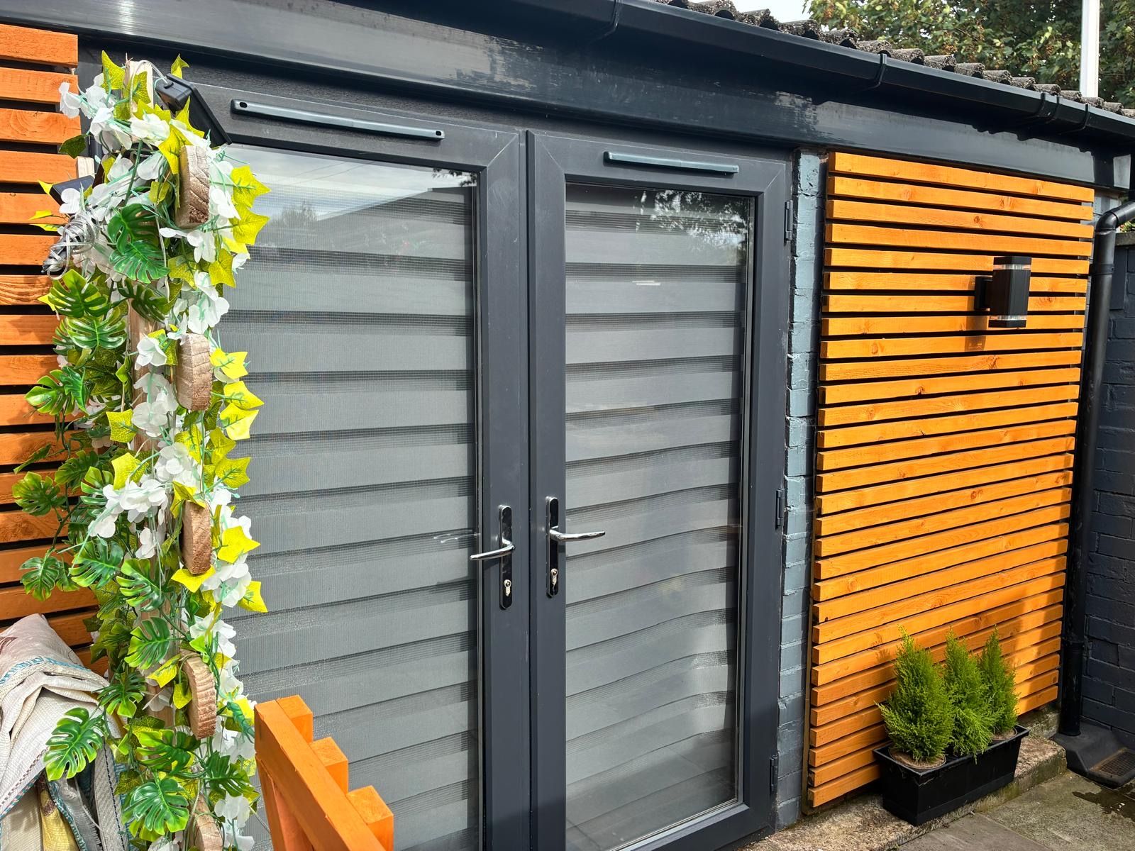 A modern building with a grey sliding door, a wooden slatted wall, and a small planter with green plants.