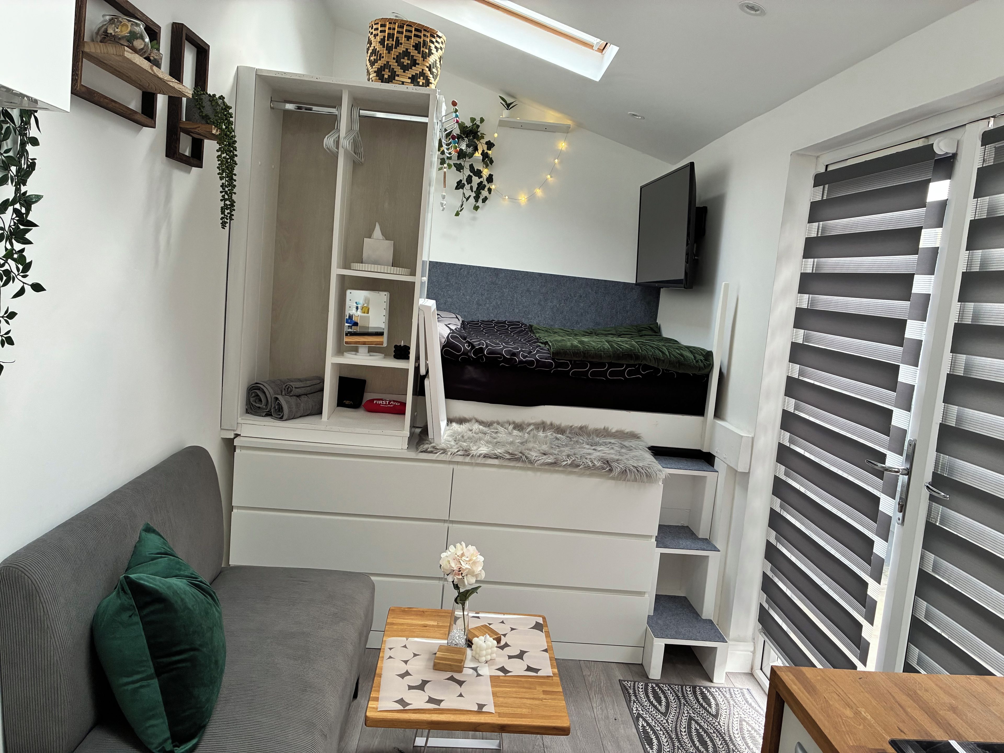 Un dormitorio pequeño con cama, armario empotrado, una mesita con un florero y un sofá gris.