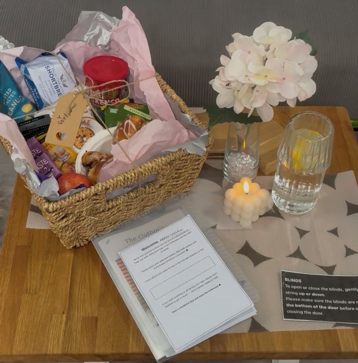 A welcome sign, a wicker basket with snacks, a candle, and a water bottle on a table.