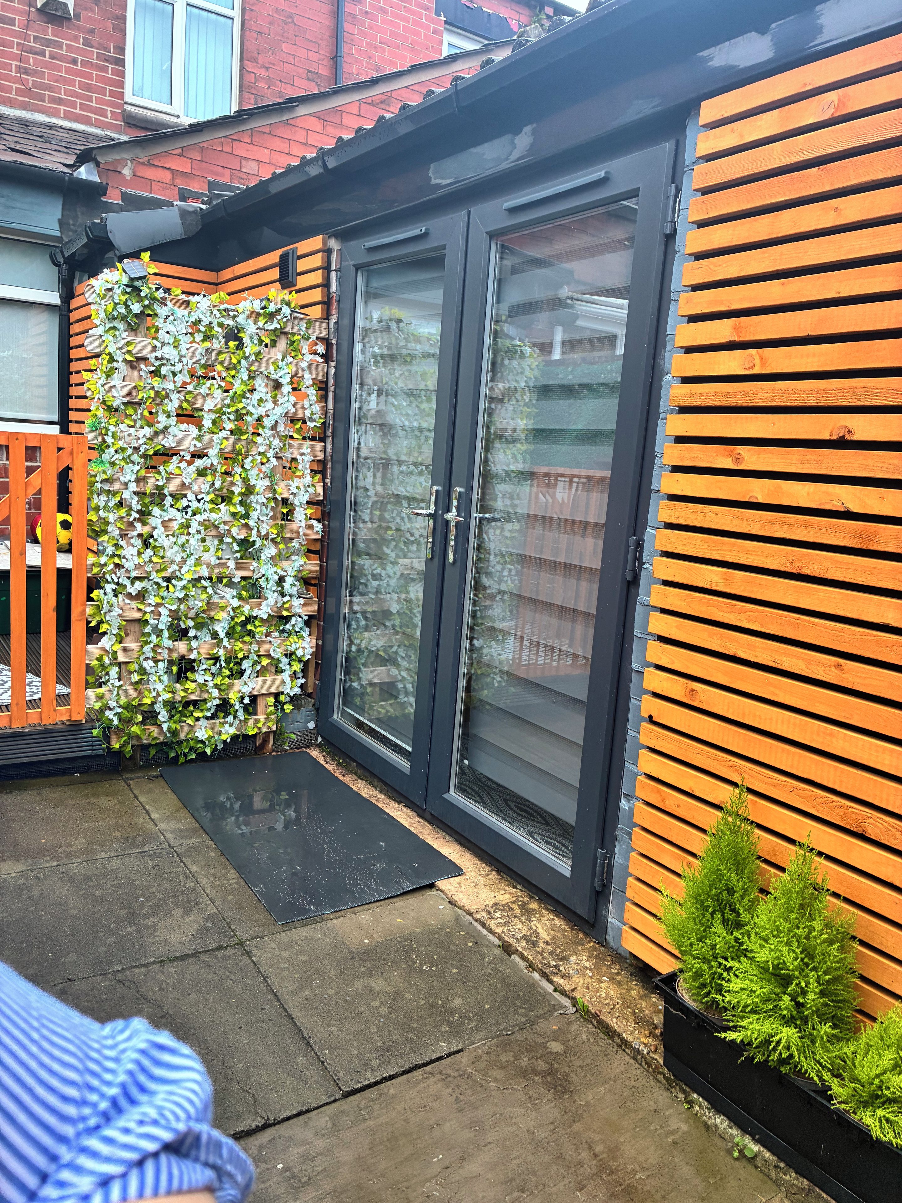 A residential property with a black sliding door, a wooden fence with climbing plants, and a paved patio area.