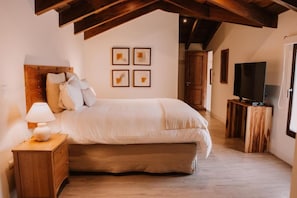 A bedroom with a bed, a nightstand with a lamp, a television on a wooden stand, and a door leading to another room. - Bärehuus Boutique Hotel (Antigua Guatemala)