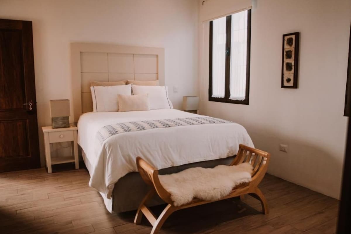 A bedroom with a bed, bedside tables, a wooden bench, a window, and a door.