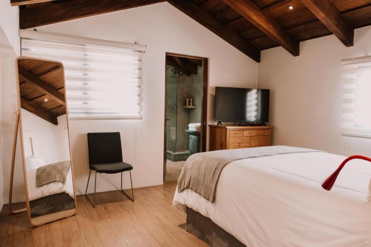 A bedroom with a bed, a chair, a television, and a wooden ceiling.