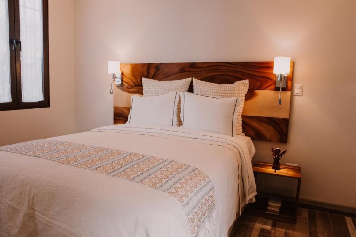 A neatly made bed with a patterned bedspread, two wall-mounted lamps, and a small bedside table with a figurine.