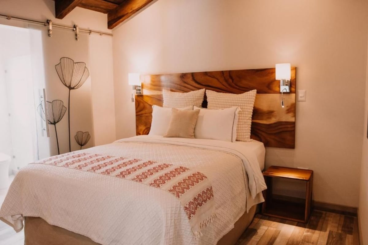 A bedroom with a wooden headboard, a bed with white bedding, a bedside table, and a wall-mounted lamp.