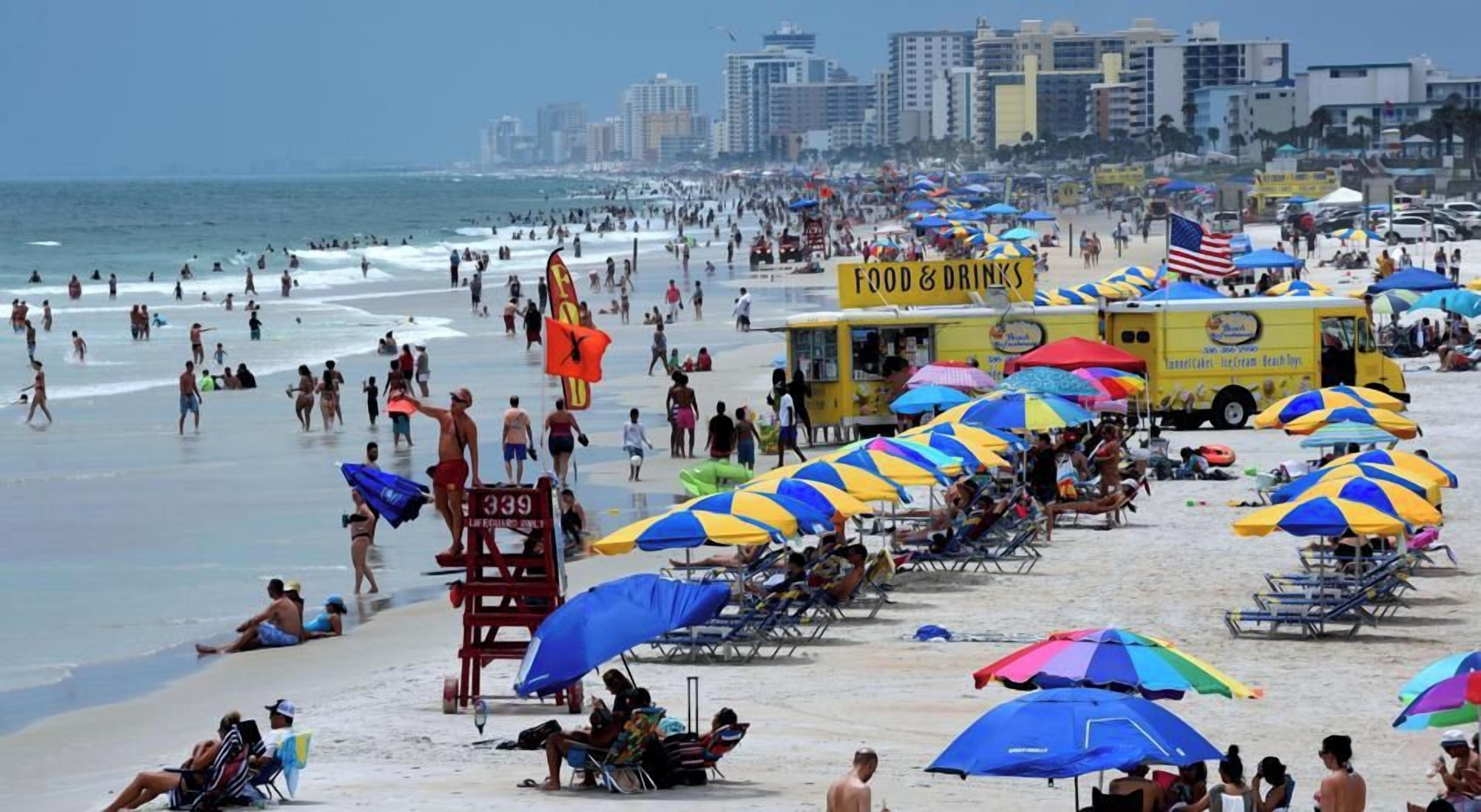 Une plage bondée, avec des parasols et des chaises de plage, une tour de surveillance pour les sauveteurs et un camion-restaurant.