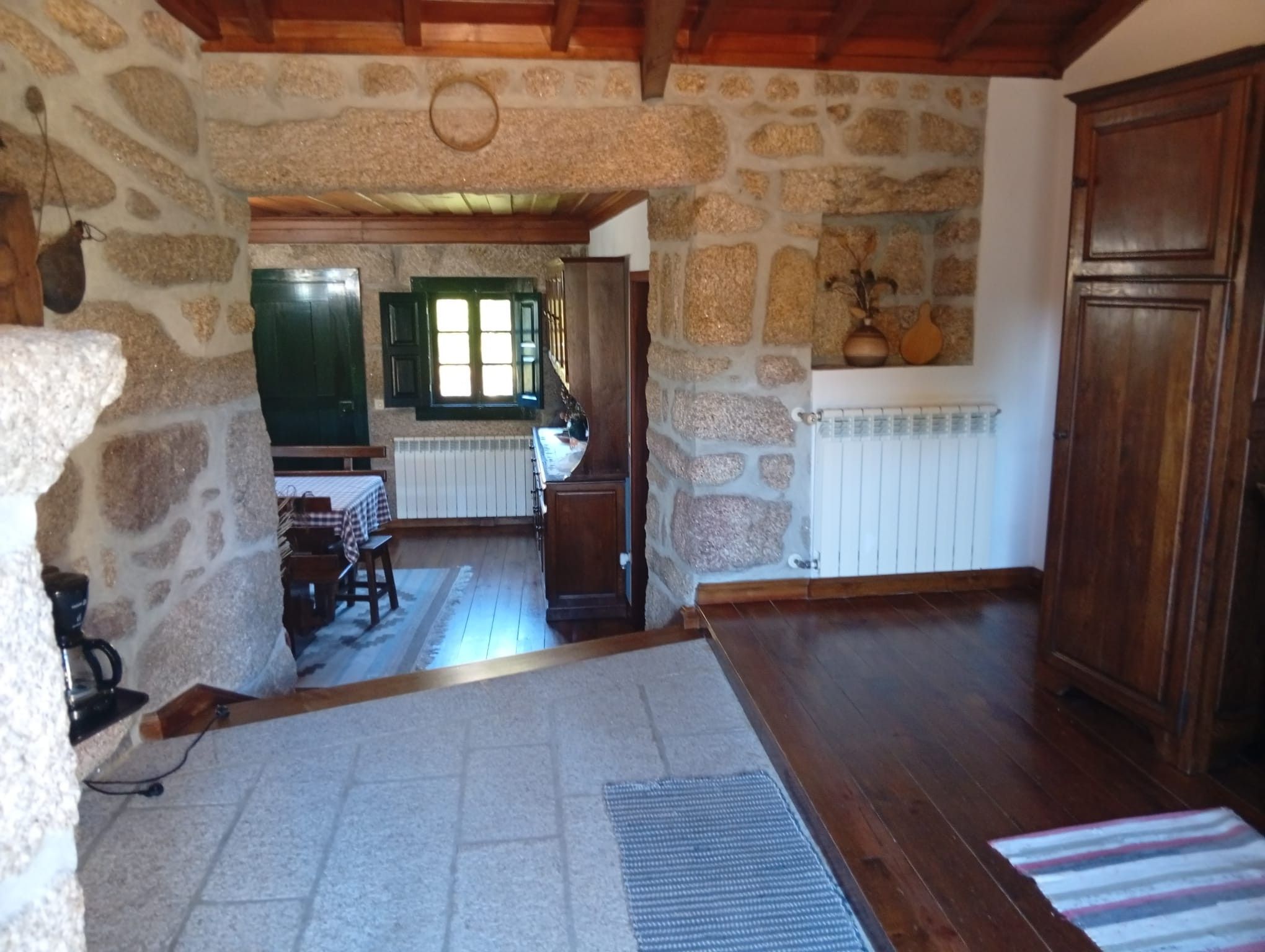 A stone-walled room with a wooden floor, a radiator, a table with chairs, and a wooden cabinet.
