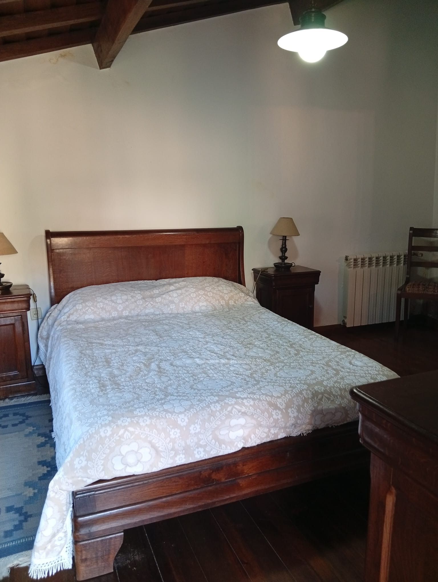 A bedroom with a wooden bed, a nightstand with a lamp, a radiator, and a chair.