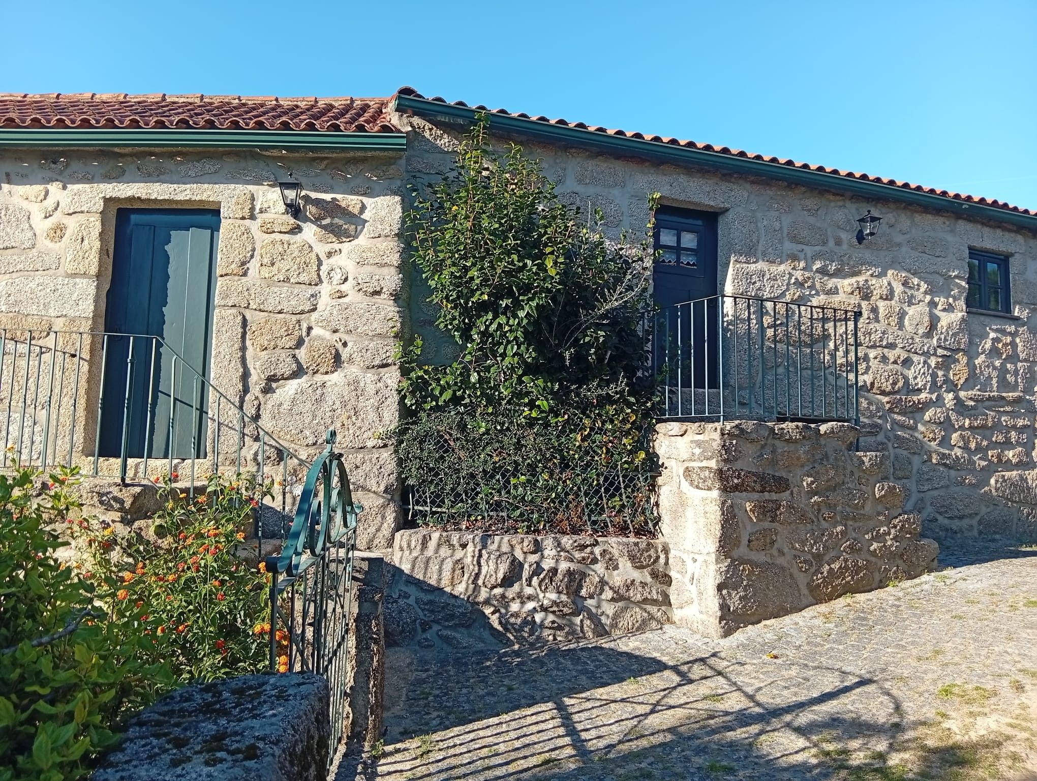 Un bâtiment en pierre avec deux fenêtres, un portail en métal et un petit jardin.