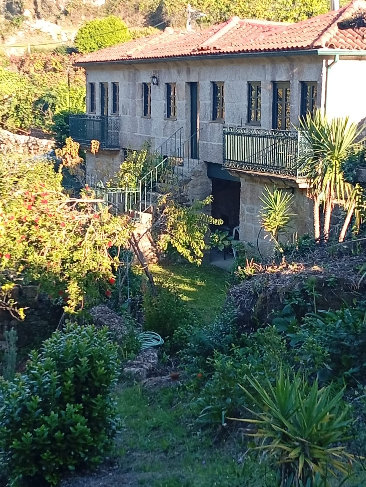A two-story stone building with a balcony, surrounded by lush greenery and a garden.