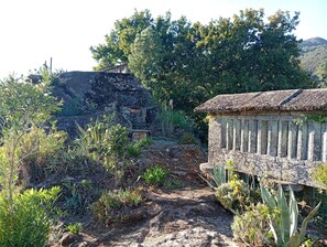 A rustic stone building with a thatched roof, surrounded by a variety of vegetation and a dirt path leading to it. - Room 'Suite 2' with Mountain View, Shared Garden and Wi-Fi (Soajo)