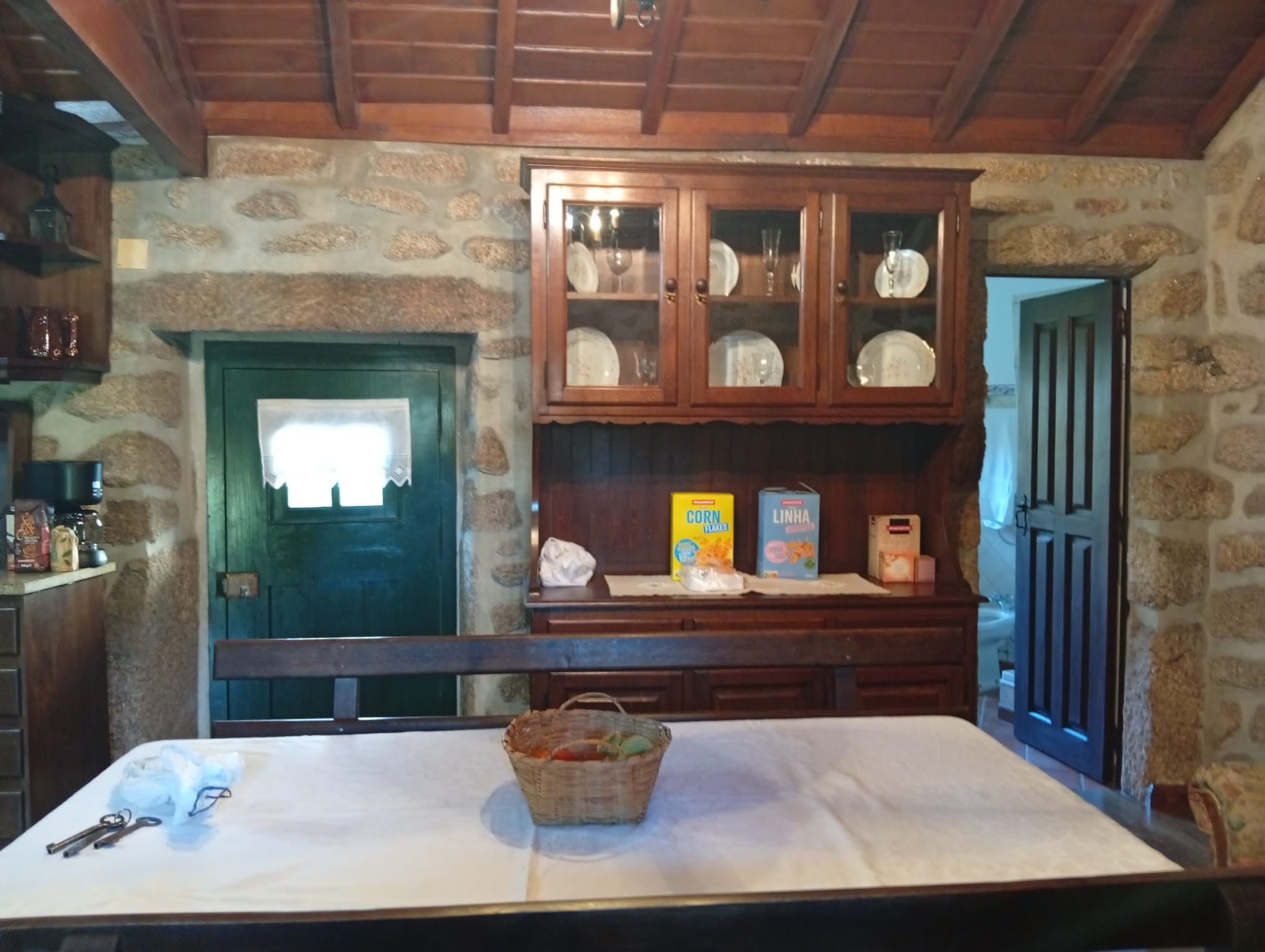 A rustic kitchen with a stone wall, wooden cabinets, and a table with a basket and cereal boxes.