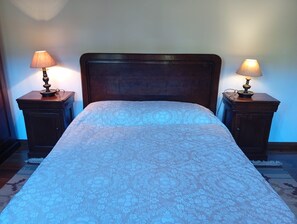 Cama com colcha estampada com motivos florais, com duas mesas de cabeceira laterais, cada uma com uma luminária, em um quarto com cabeceira de madeira.