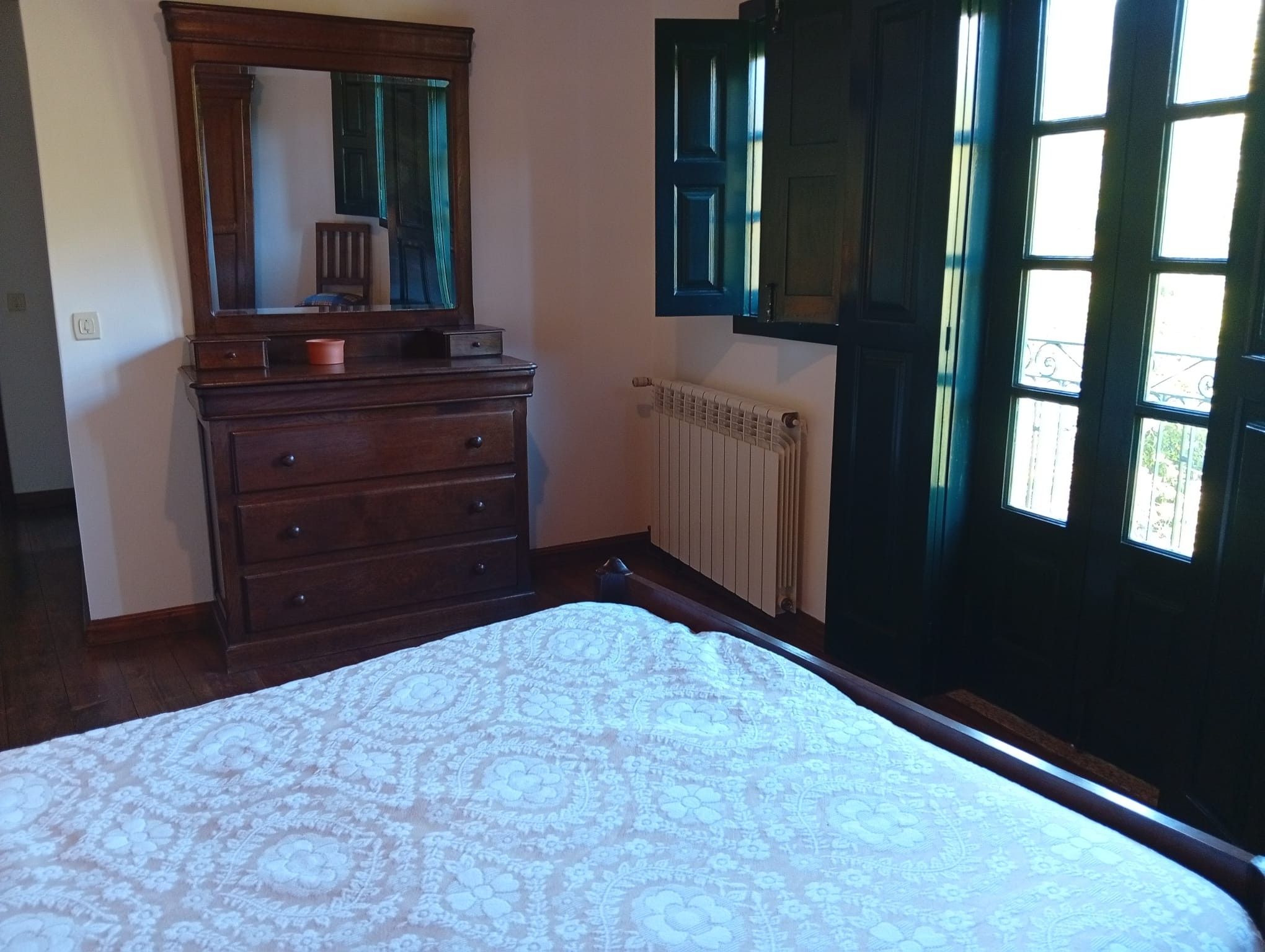 Quarto com cômoda de madeira, cama grande com colcha estampada, espelho sobre a cômoda e janela com persianas.