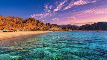 A coastal landscape with clear turquoise waters, a sandy beach, and mountainous terrain in the background.
