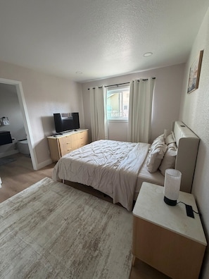 A bedroom with a bed, bedside table, TV, and a window with curtains. - Brand-New Renovated 4BR/2BA l Prime Hayward Location (Hayward)