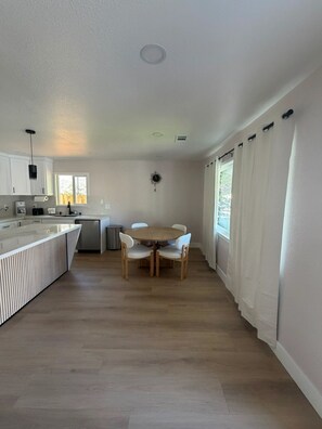 A modern kitchen with white cabinetry, a wooden dining table with chairs, and a window with curtains. - Brand-New Renovated 4BR/2BA l Prime Hayward Location (Hayward)
