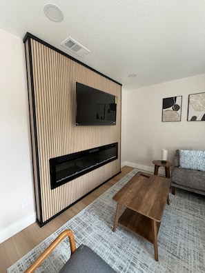 A modern living room with a flat-screen TV mounted on a wooden panel wall, a fireplace below, a grey sofa, a wooden coffee table, and a patterned rug. - Brand-New Renovated 4BR/2BA l Prime Hayward Location (Hayward)