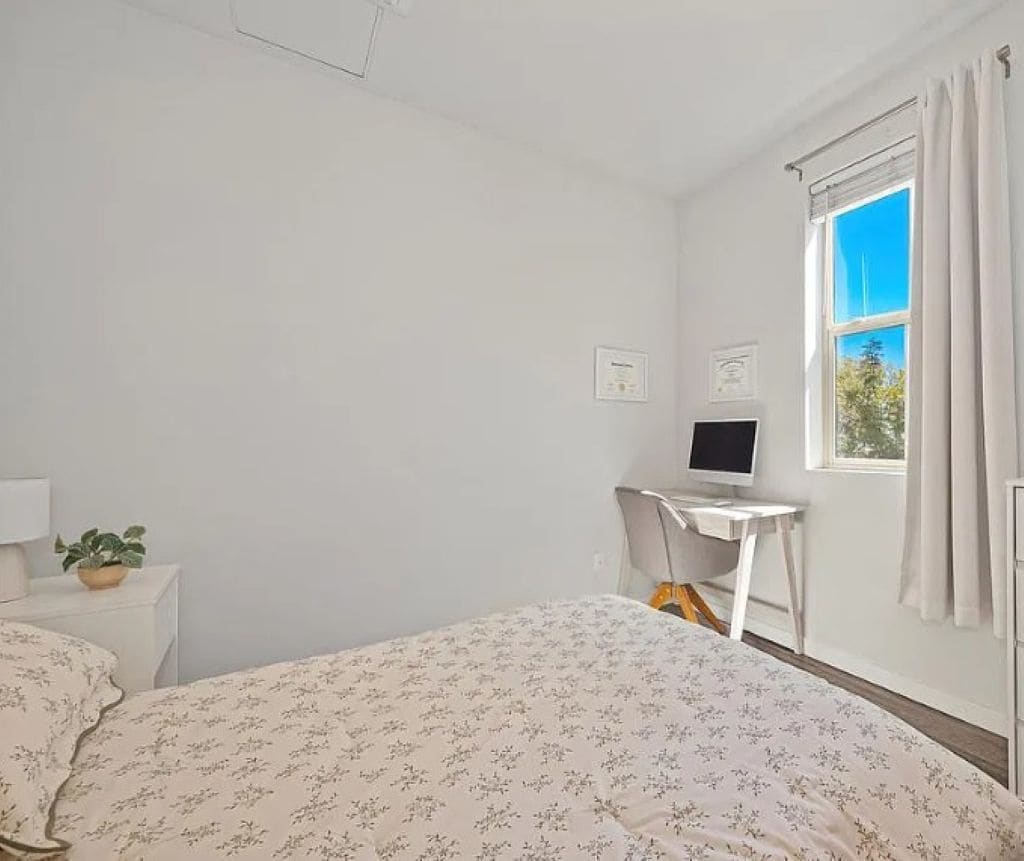 A bedroom with a bed, a desk with a computer, a window with curtains, and a potted plant.