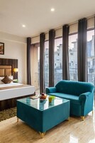 Una moderna camera d'albergo con un letto, un divano color petrolio, un tavolino in vetro e ampie finestre con vista sulla città.