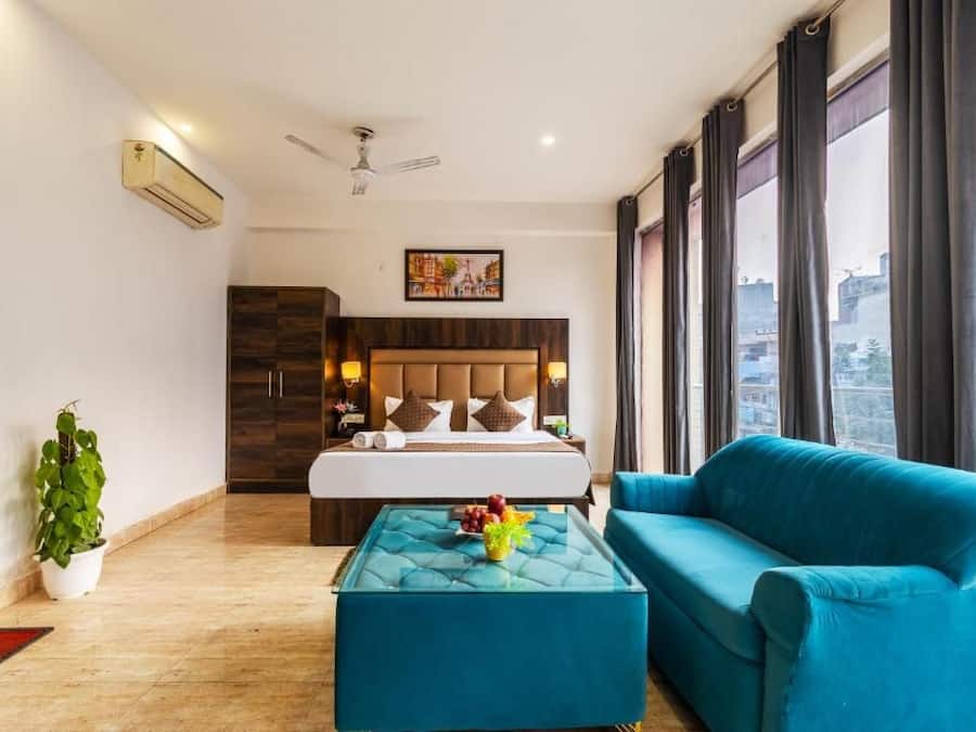 A hotel room with a bed, a blue sofa, a glass coffee table with fruit, a potted plant, and a city view.