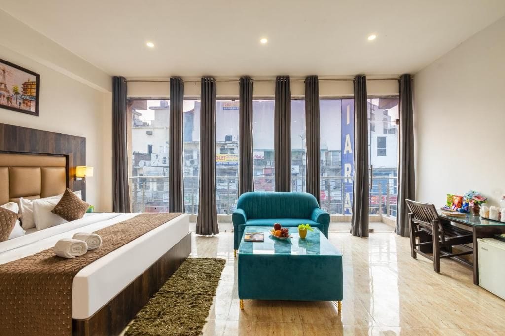 Habitación de hotel con una cama, un sillón azul, una mesita, un escritorio y vistas a la ciudad a través de amplias ventanas.