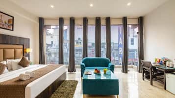 Habitación de hotel con una cama, un sillón azul, una mesita, un escritorio y vistas a la ciudad a través de amplias ventanas.