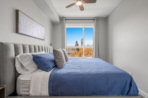 A bedroom with a bed, a ceiling fan, a window with a view of trees, and a painting on the wall. - `Auburn` 1 King  1 Queen Bed | UG Parking | Sofa Bed (Calgary)
