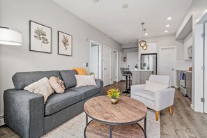 Una sala moderna con un sofá gris, una mesa de centro redonda de madera y un sillón blanco. Hay una zona de cocina con un refrigerador de acero inoxidable y una mesa de comedor.