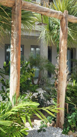 Een pergola van hout met een waterornament, omgeven door diverse tropische planten en een gebouw op de achtergrond.