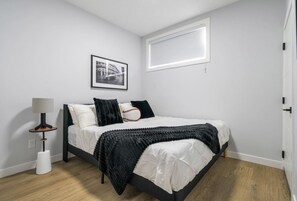 A bedroom with a bed, a bedside lamp, a framed picture on the wall, and a window. - `Boston` King Bed | Private Suite + 7 Min to DT (Calgary)