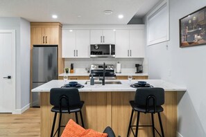 A modern kitchen with white cabinets, a stainless steel refrigerator, a microwave, and a coffee maker. There are two black chairs at the island. - `Boston` King Bed | Private Suite + 7 Min to DT (Calgary)