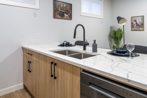 A modern kitchen with a white marble countertop, two sinks, wooden cabinets, and a dishwasher. - `Boston` King Bed | Private Suite + 7 Min to DT (Calgary)