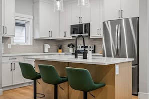 Eine moderne Küche mit weißen Schränken, einem Edelstahlkühlschrank, einer zentralen Kochinsel mit Barhocker und einer Mikrowelle.