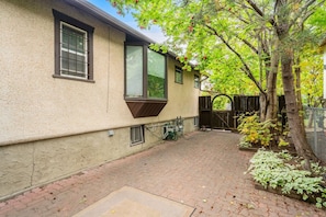 A house with a paved backyard, a window with a brown awning, and a tree with green leaves. - The Woodside | 2 King | Foosball | Pull out Couch (Calgary)