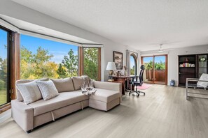 A modern living room with a sectional sofa, a desk, and a chair, large windows with a view of trees, and a sliding glass door leading to an outdoor area. - The Woodside | 2 King | Foosball | Pull out Couch (Calgary)