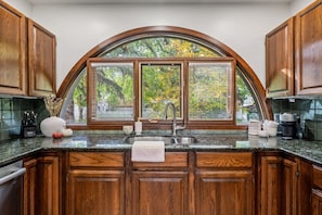 A kitchen with wooden cabinets, a granite countertop, a large window with blinds, and a stainless steel sink. - The Woodside | 2 King | Foosball | Pull out Couch (Calgary)