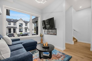 A modern living room with a sofa, a TV mounted on the wall, and a view of the outside. - ‘Indigo’ 3 Bedroom | Garage | King + 7 Min to DT (Calgary)