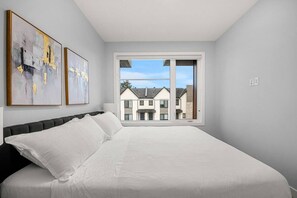 A bedroom with a large bed, a window with a view of houses, and two abstract paintings on the wall. - ‘Indigo’ 3 Bedroom | Garage | King + 7 Min to DT (Calgary)