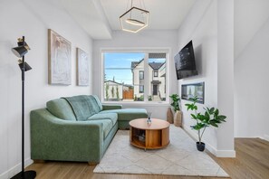 A modern living room with a green sofa, a round wooden coffee table, and a large window offering a view of a house. - `The Juniper` 3 Bed | King | Garage | 5 Min to DT (Calgary)
