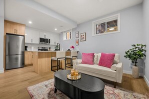 A modern kitchen with white cabinets, a stainless steel refrigerator, and a microwave. There is a dining area with a white sofa and a black coffee table. - `Paris` King Bed | 7 Mins to DT + Free Parking (Calgary)