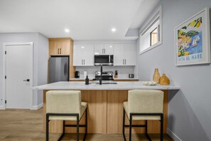 A modern kitchen with white cabinets, a central island with bar stools, and a painting on the wall. - `Greece` King Bed | Private Suite + 7 Min to DT (Calgary)