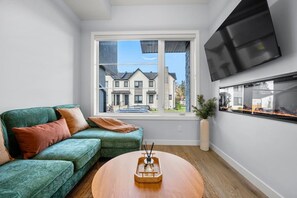 Ett vardagsrum med en grön soffa, ett soffbord, en tv som är monterad på väggen och ett fönster med utsikt över ett bostadsområde.