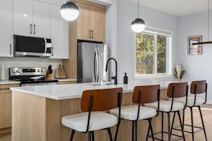 A modern kitchen with a central island, bar stools, stainless steel appliances, and pendant lighting. - `Jade` 3 King Bedrooms | Garage + 7 Min to DT (Calgary)