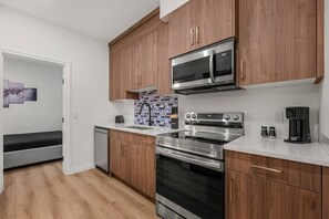 A compact kitchen with wooden cabinets, a stainless steel microwave, an oven, and a coffee maker. - The Nook | 2 Bedroom | Close to DT Hospital & UofC (Calgary)