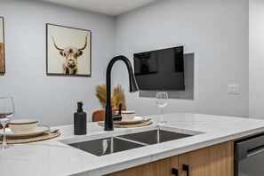 A modern kitchen with a double sink, a black faucet, a mounted flat-screen TV, and a framed artwork of a bull's head. - `Stampede` King Bed | 7 Min to Downtown + Parking (Calgary)